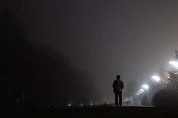 A man standing with his back to us in a jacket on an evening alley in the fog illuminated by evening white lamps against a background of birches and shrubs.