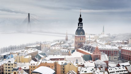 Panoramic aerial view of the Riga old town and Daugava river from St. Peter's Church in winter. Morning fog, snow-covered cable-stayed bridge, cathedral, houses. Latvia. Landmarks, sightseeing - obrazy, fototapety, plakaty