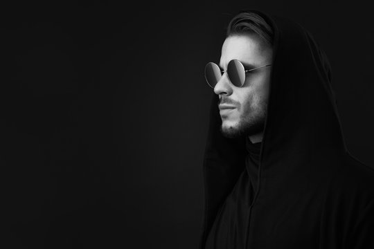 Young Man With Beard In Hoody And Glasses. Black And White Portrait Of Guy In Cape On Dark Backgound. High Fashion Model Posing In Studio. Free Space For Text.