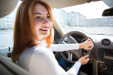 Young redhead woman driver behind a wheel driving a car smiling happily.