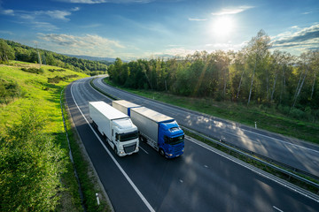 Blue truck overtaking white truck on an asphalt highway between forests under radiant sun. View...
