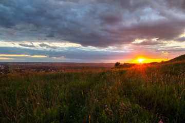 Naklejka na ściany i meble Sunset in dramatic cloudscape over wild herb field with distant city in view.