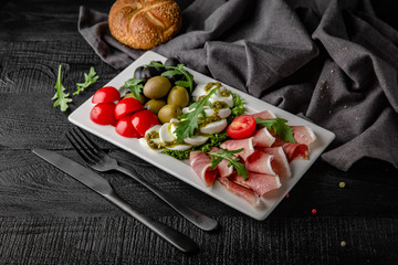 Caprese appetizer: mozzarella with pesto, cherry tomatoes, arugula, green olives, bacon, prosciutto and black olives. On a black background