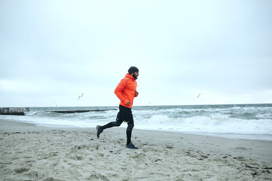 Full-length photo of young bearded sportsman dressed in athletic clothes and warm orange coat working out and running by seaside on gray cold weather. Fitness male model