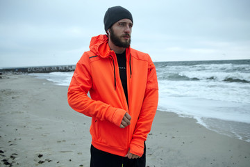 Outdoor shot of young pensive bearded male looking thoughtfully ahaead while fastening zipper on...