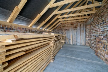 Stack of wooden boards in unfinished brick house with concrete floor, bare walls and wooden roofing frame attic under construction.