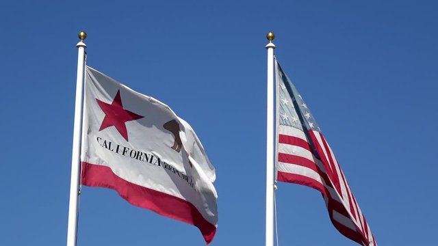 4K. American and Californian flags waving in the wind on flagpole in the city of Los Angeles with clear sky and cloudy background. Close up of a cloth sill flags fluttering through the air. 