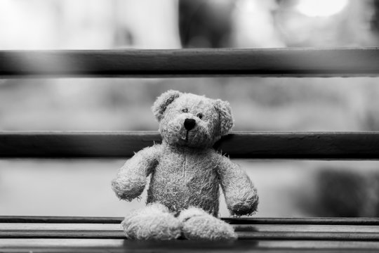 Black and White photo of Teddy bear with sad face sitting on wooden beance , Lonely teddy bear sitting alone outside in gloomy day, Lonely concept, International missing children's day.
