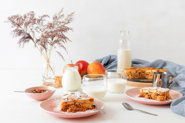 White wooden table with apple bread and butter pudding, jar with sour cream, glass of milk, background decorated with few apples and vase with dry grass.