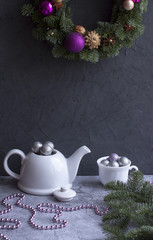 A cozy Christmas composition with a white teapot filled with shiny silver Christmas balls and a white Cup filled with balls against a dark wall and a hanging Christmas wreath of Nobilis with pink ball