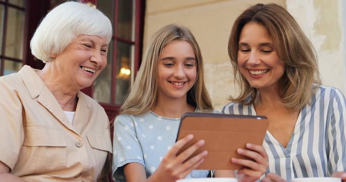 Beautiful and cute Caucasian teenage girl standing outside with her cheerful mother and grandmother and showing them photos or video on the tablet device.