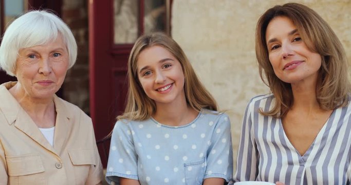 Portrait shot of the cute and pretty Caucasian teen girl drinking tea and smiling cheerfully to the camera with her grandmother and mother at the cafe terrace outdoor.