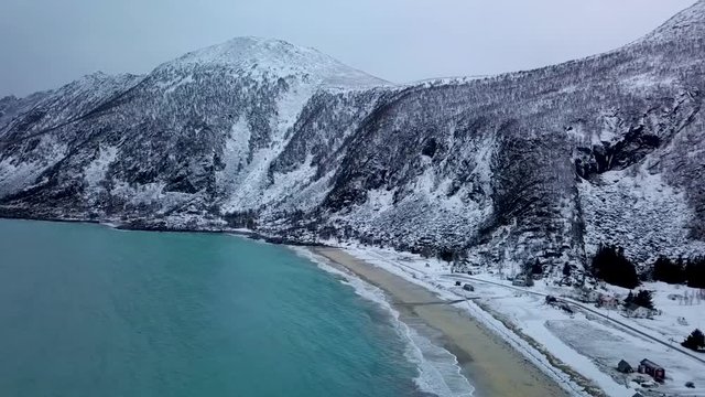 flying over drone of a snowy Norway beach. No sun. Turquoise blue water and snow on the surrounding mountains