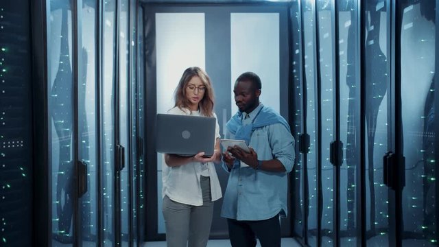 Two multi-ethnic IT workers cooperating in data server cabinet. Male and female server specialists shrugging shoulders hesitating or having problem with connection. Data center team.