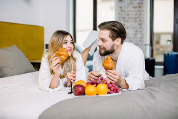 Enjoying breakfast, fruits, croissants, champagne. Man and woman wearing white bathrobes enjoying breakfast in hotel room