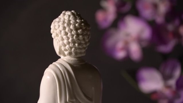 White Buddha figurine rotates on a black grange background with orchid flower