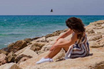 Fototapeta na wymiar Woman natural beauty without retouching. Pretty girl sit on stones on beach at sea coast enjoying pleasant weather on a sunny day. Travel, relax, lifestyle. Sharpness at sea. Girl out of focus