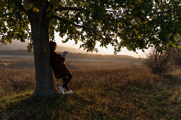 Young girl in red sweater leaned on a big tree among the meadow and holds book in hands, enjoying sunny autumn day