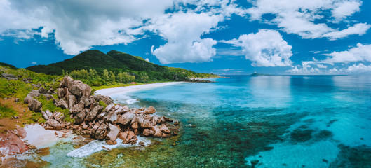 Aerial panoramic view of impressive Grand Anse beach on La Digue island in Seychelles. Granite boulders and turquoise ocean lagoon with white clouds above - obrazy, fototapety, plakaty