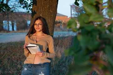 Close up portrait of a beautiful brunette girl in glasses that leaned on big tree with book in hands