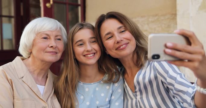 Caucasian beautiful smiled and happy woman taking selfie photo on the smartphone camera with her teen daughter and senior mother outside.