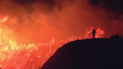Slow motion shot, of a silhouette man standing on a hill, in front of burning nature of the Californian wildfires, at night time, in Northridge, Los Angeles, California, USA