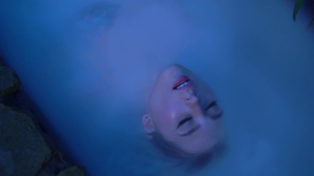 Portrait of gorgeous attractive brunette girl with elegant waterproof makeup. Woman in old stone bath with milk and steam on nature at evening dusk. Fashion shooting, sexy girl dancing in white water