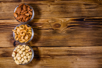 Fototapeta premium Various nuts (almond, cashew, hazelnut) in glass bowls on a wooden table. Vegetarian meal. Healthy eating concept. Top view