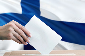 Finland election concept. Side view woman putting a ballot in a box on national flag background.