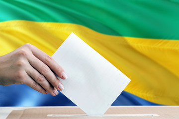 Gabon election concept. Side view woman putting a ballot in a box on national flag background.