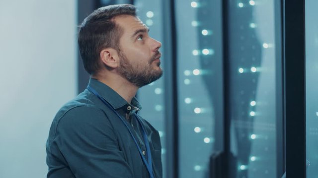 Bearded IT expert examining server cabinet with notebook, doing web hosting maintenance operation on supercomputer in cyber workspace of data center.