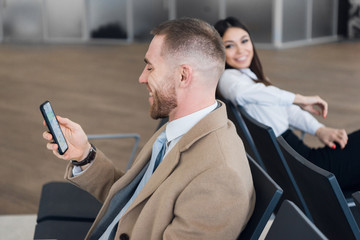 Fototapeta na wymiar Young businessman and woman waiting at airport lounge. Man looking at phone. Business travelers waiting for their flight at airport terminal.