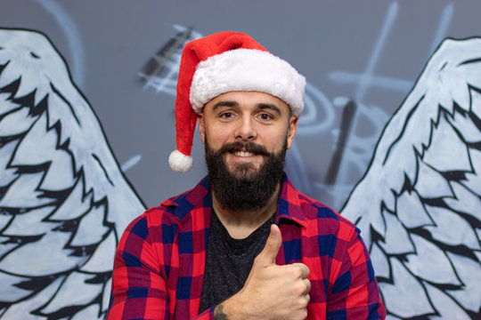 Dark Bearded Man With A Santa Claus Red Hat Posing In Front Of Angel Wings Wall Graffiti With His Tumbs Up. Christmas Holidays Concept.