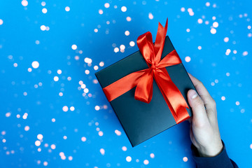Right mans hand gifts a present black box with red ribbon on classic blue background, the color of the year 2020. Enough place for text. Bokeh style.