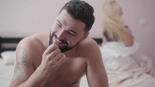 Portrait of young Caucasian bearded man looking back at satisfied blond woman sitting on bed at the background. Smiling guy resting in bedroom after sex.