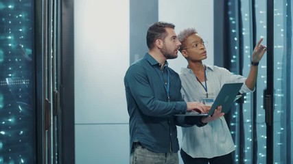 Mixed-race professional IT team cooperating in data center,. Attractive male and female colleagues doing deep diagnostics of server cabinet with laptop technology. - Powered by Adobe