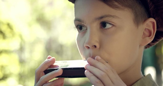 Close up of the nice and cute small Caucasian boy in a hat playing on the musical instrument: mouth organ or harmonica. Outside.