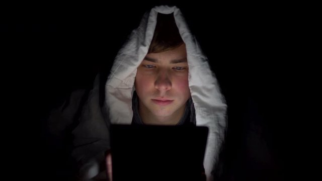 A young man lies under a blanket with a tablet in him hands. A man in the dark uses a pad. Modern electronic tablet. Close-up