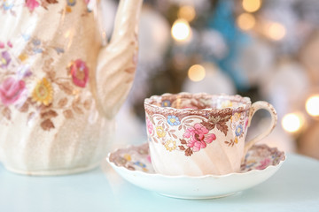Steam streams out of a colorful English tea cup on a teal table in front of a decorated Christmas holiday tree
