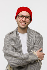 Studio portrait of cheerful stylish hipster guy with light bristles on the face smiling at camera, wearing glasses, red beanie and wrapped in plaid, arms crossed. Isolated on white background. 