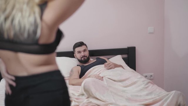 Back view of half naked blond Caucasian woman in black clothes standing in front of bearded man lying at the background. Focus changes to smiling guy waiting for his girlfriend or wife in bed.