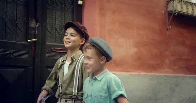 Two Caucasian small friendly boys in hats and vibtage style walking the street with a tire and talking. Retro style shooting. Outside.