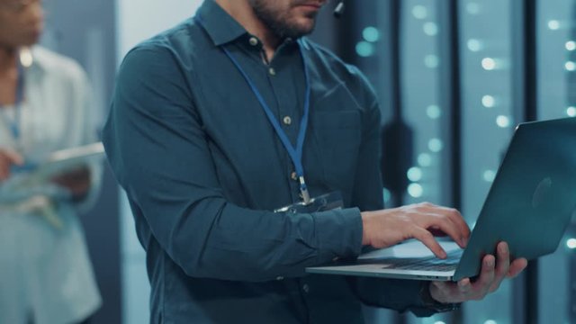 Male technician running secure tech backups on laptop inside server cabinet. Positive system support engineer smiling at camera satisfied of work in modern data center.