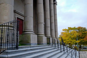 Large concrete columns on the front exterior wall of a court house. The sky is blue and cloudy in the background with a yellow tree.