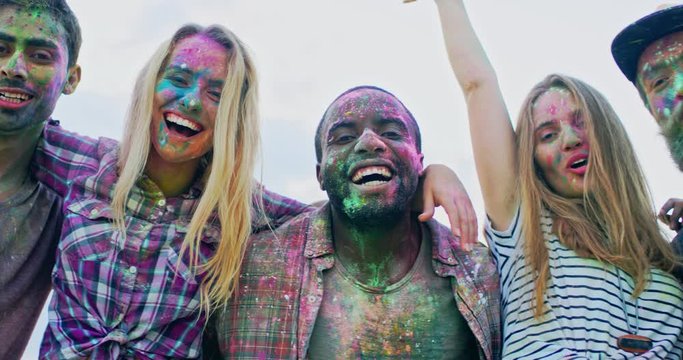 Close up of the young mixed-races cheerful friends in colorful paints during holi festival hugging and laughing to the camera. Outdoor. Portrait.