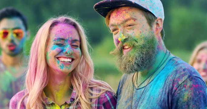 Portrait of the young Caucasian smiled and cheerful man and woman posing to the camera at the holi festival outdoors while somebody throwing colorful paints in powder on them. Close up.