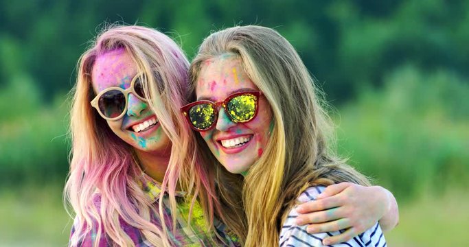 Portrait shot of the cute and beautiful Caucasian blonde girls in sunglasses smiling and laughing to the camera while posing at the holi fest celebration. Outdoor.