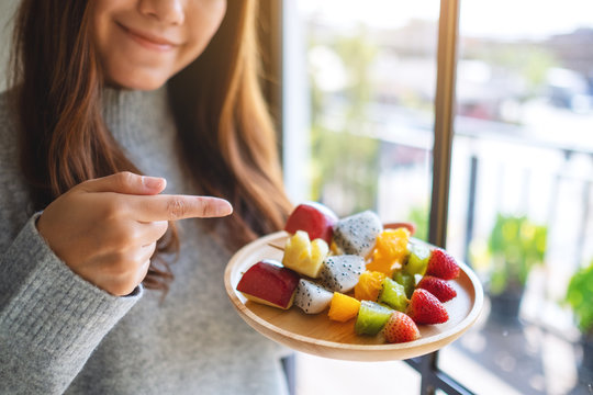 Closeup image of an asian woman holding and pointing finger at a wooden plate of fresh mixed fruits on skewers