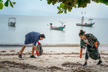 Problems of environmental pollution and oceans, a young couple in orange gloves cleans plastic and garbage in a black trash bag on a tropical beach in Thailand.