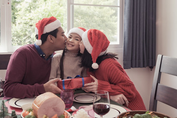 Asian family daughter hold gift with father and mother kissing her on Christmas day, they are happy and have meal together in dining room that decorated with Christmas theme